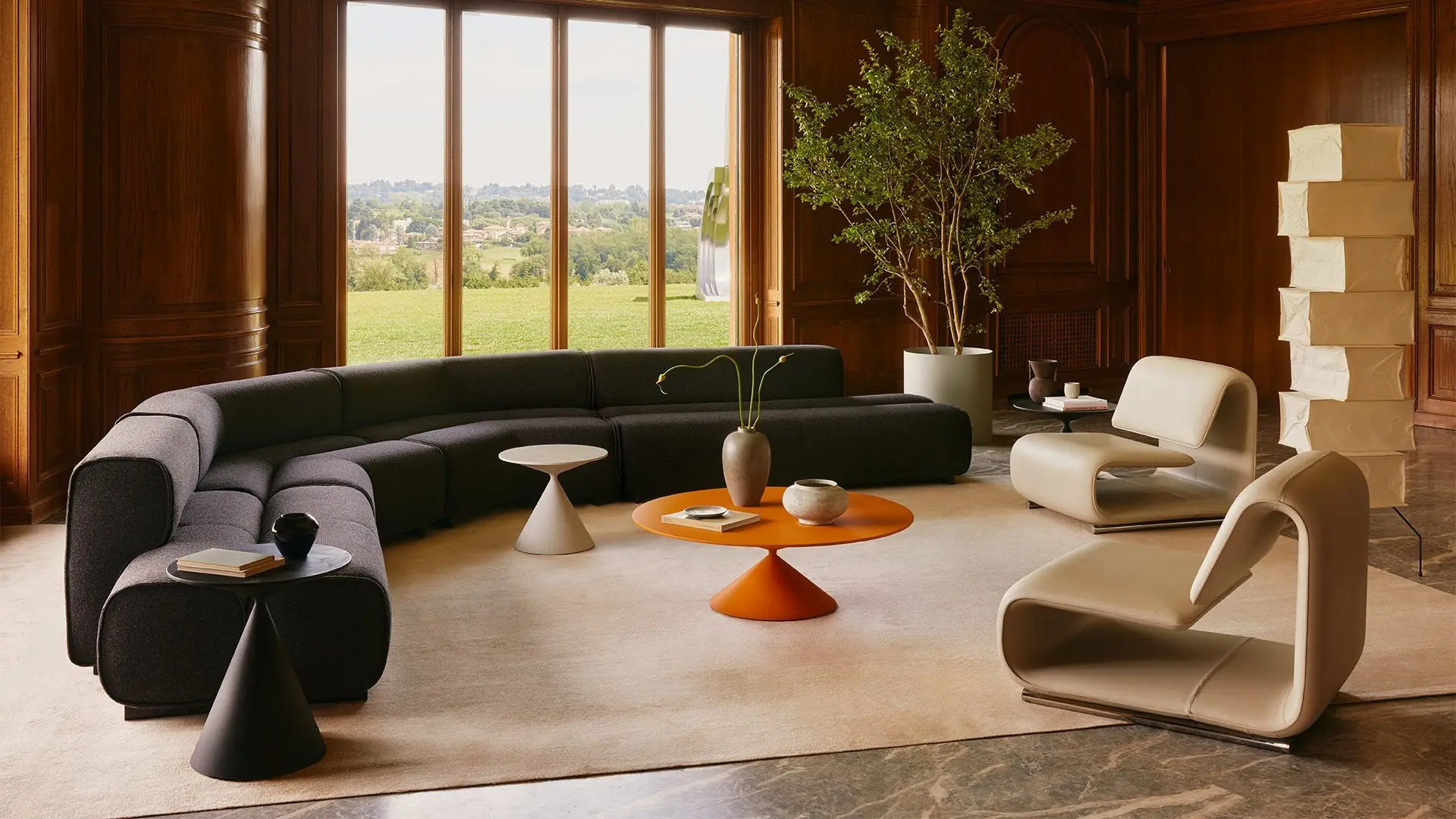 Curved black sofa, white lounge chair and an orange coffee table in a timber-panelled room with a view of hills