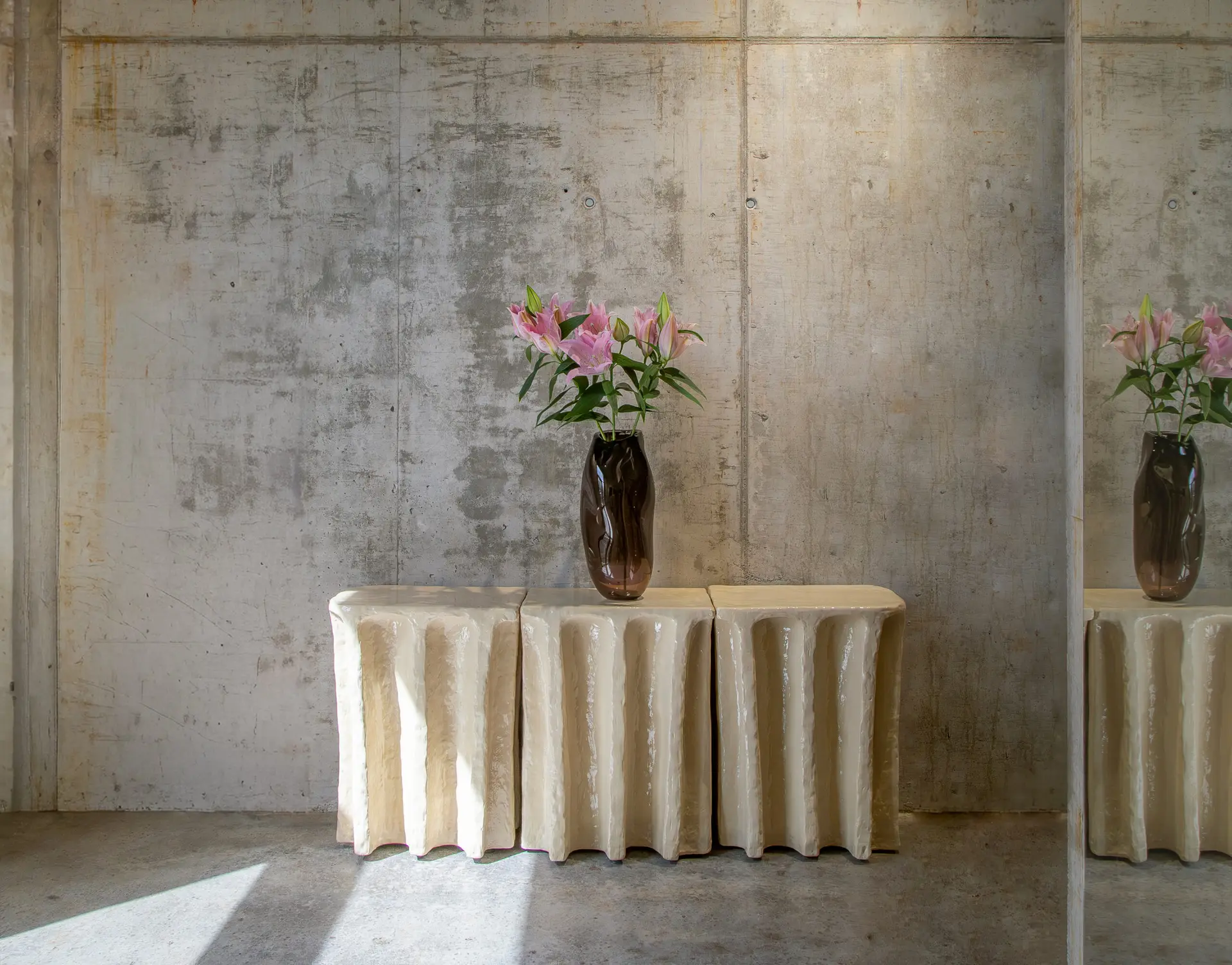 Sculptural cream consoles with draped legs and vases of lilies against a concrete wall