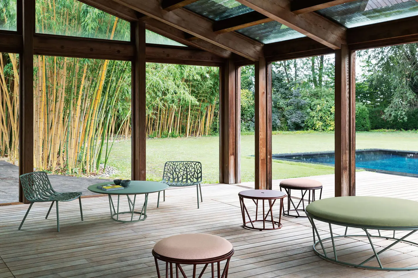 Aluminium lounge chairs, side tables and a round pouf under a timber pergola beside a pool