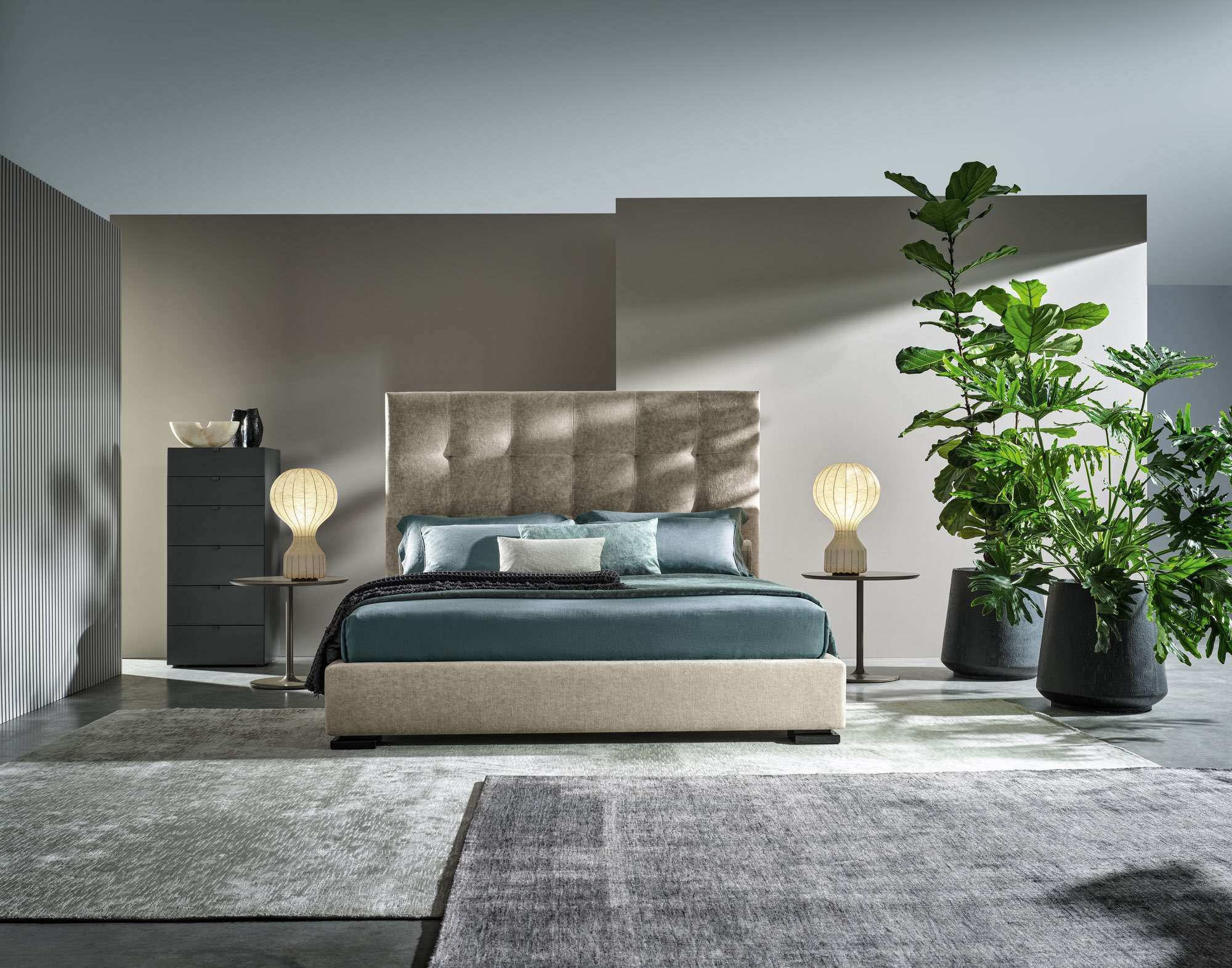 Bed with a quilted fabric headboard and teal bedding between two glowing bedside lamps, with tall plants in dark pots