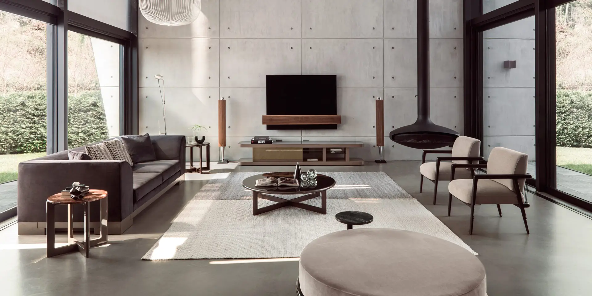 Living room with a grey sofa, two pale armchairs and a round ottoman before a concrete wall and tall glass walls