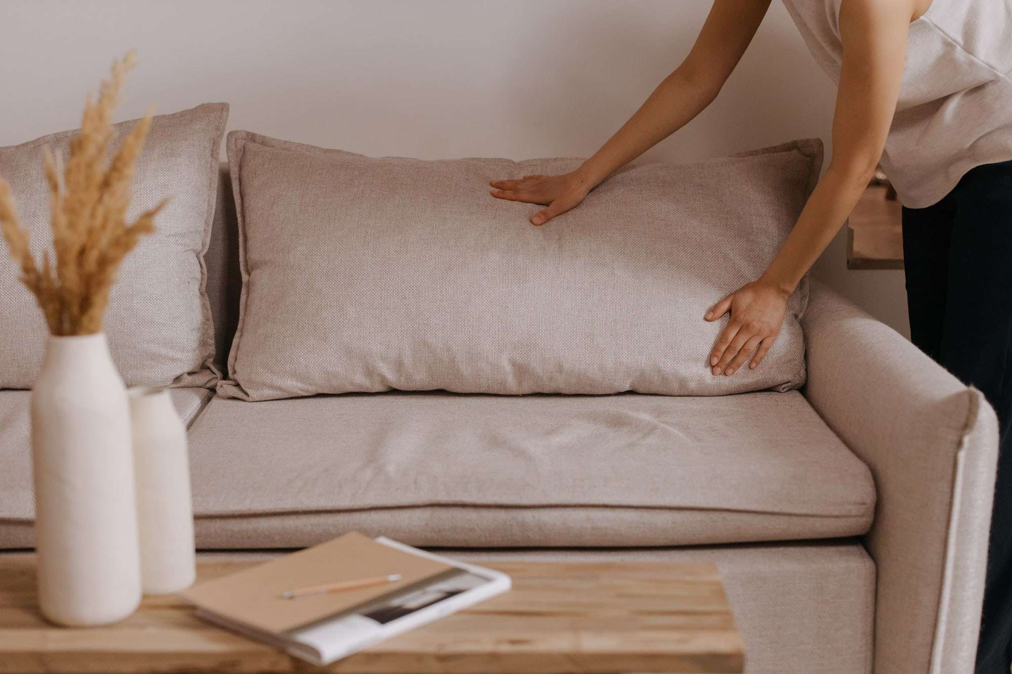 Hands setting a cushion on a pale linen sofa beside a vase of dried grasses