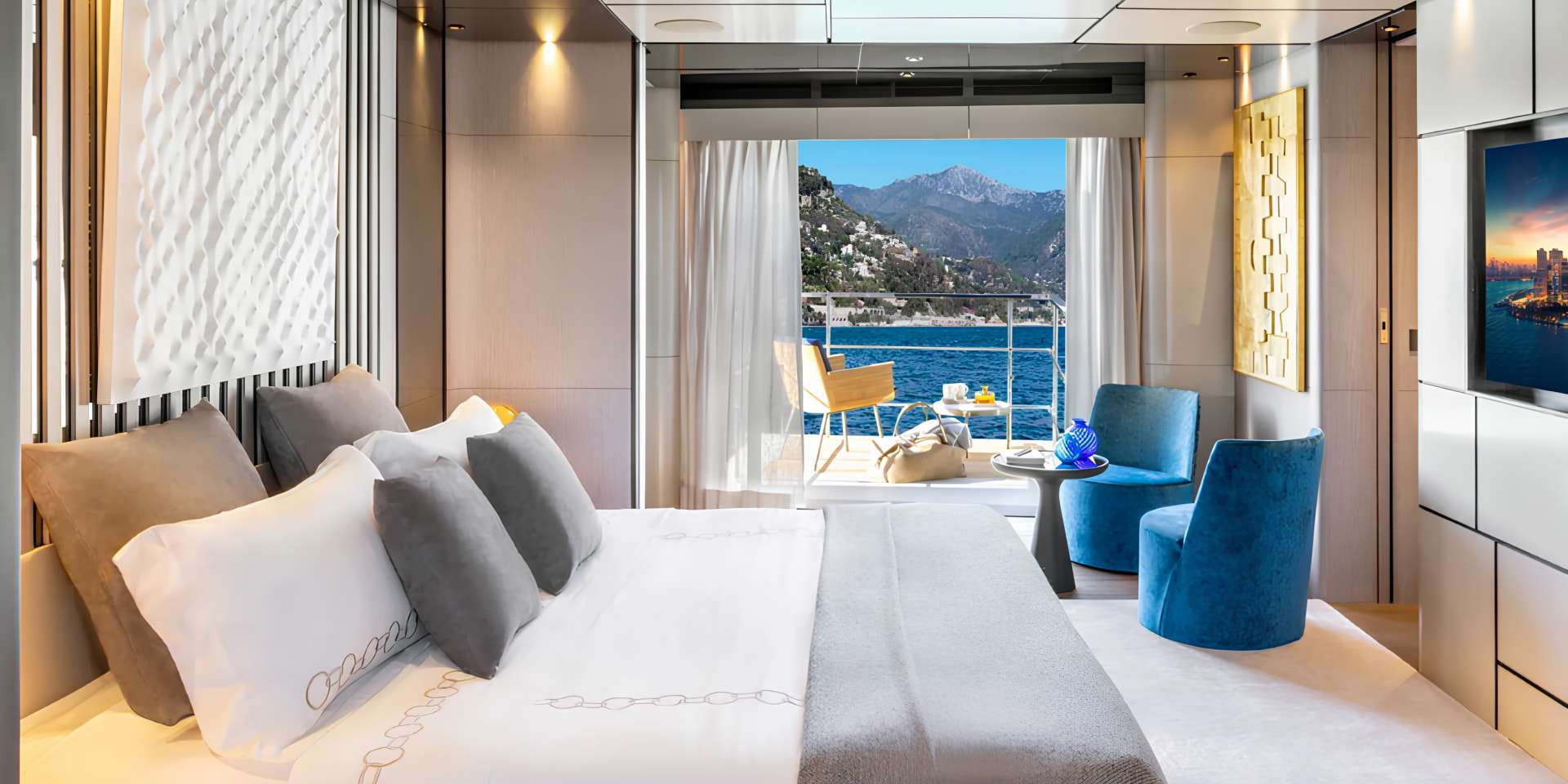 Bedroom with a white bed and blue swivel armchairs opening onto a balcony over the sea