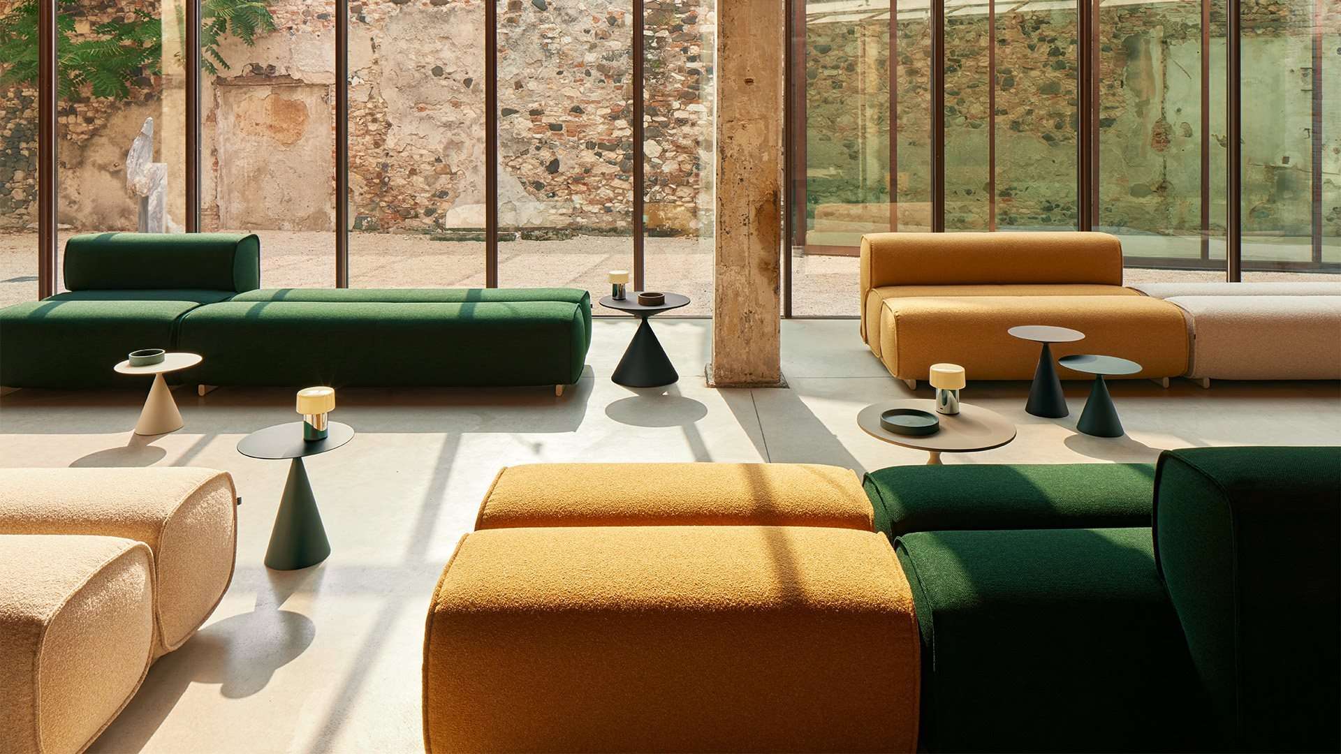 Green and ochre modular sofas with small cone tables in a glass room off a stone courtyard