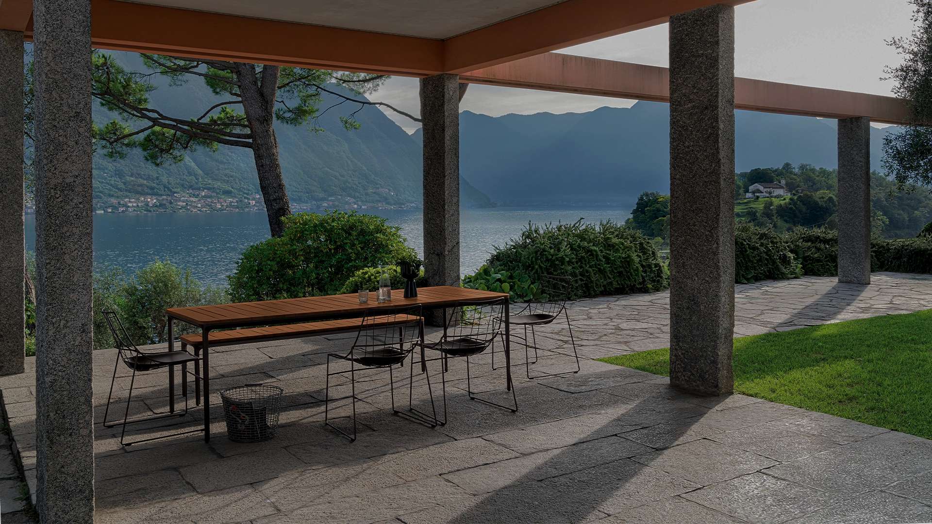 Outdoor dining table and wire chairs under a loggia overlooking a lake