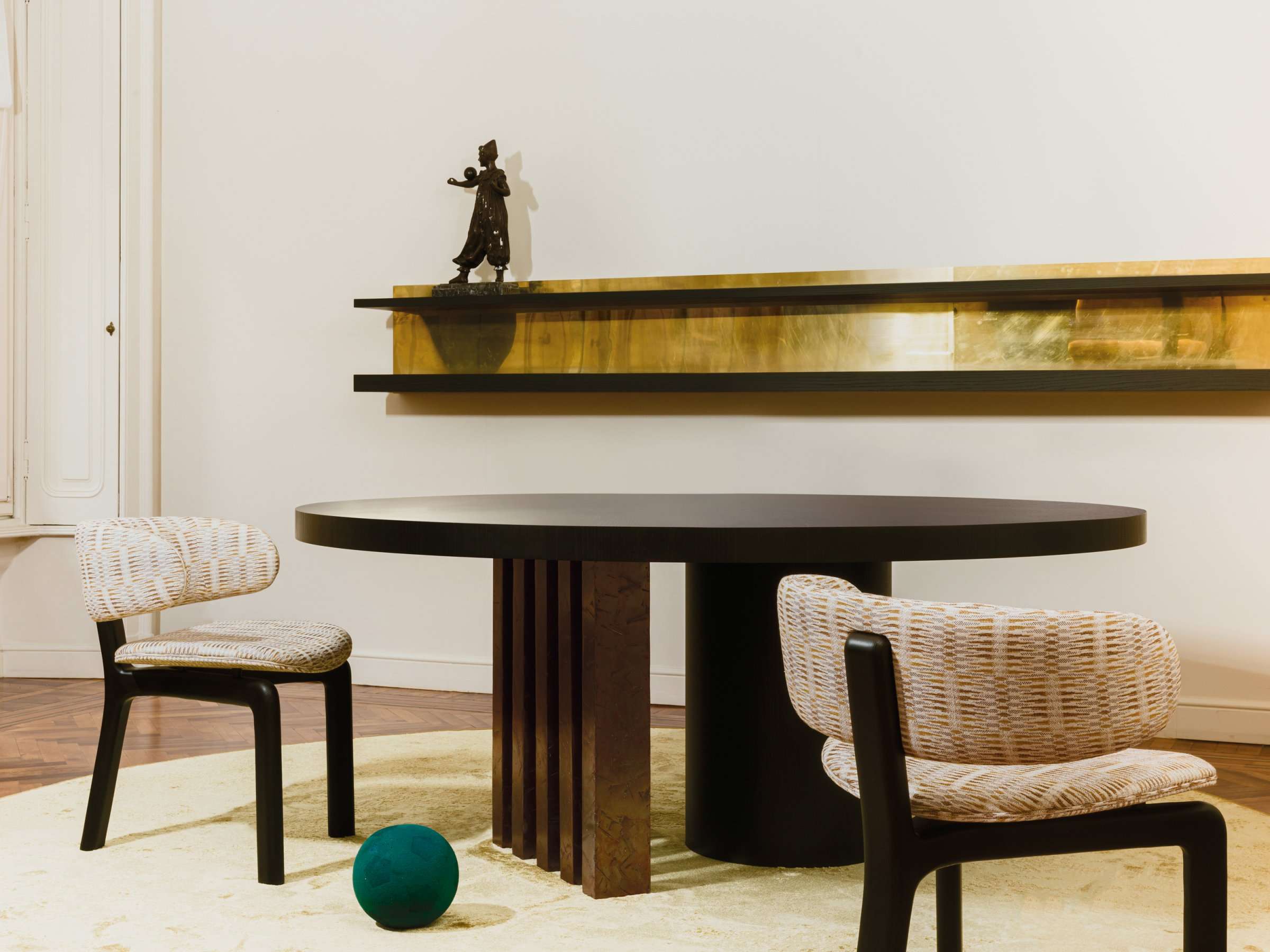 Round dark dining table on a fluted base with upholstered chairs below a brass shelf