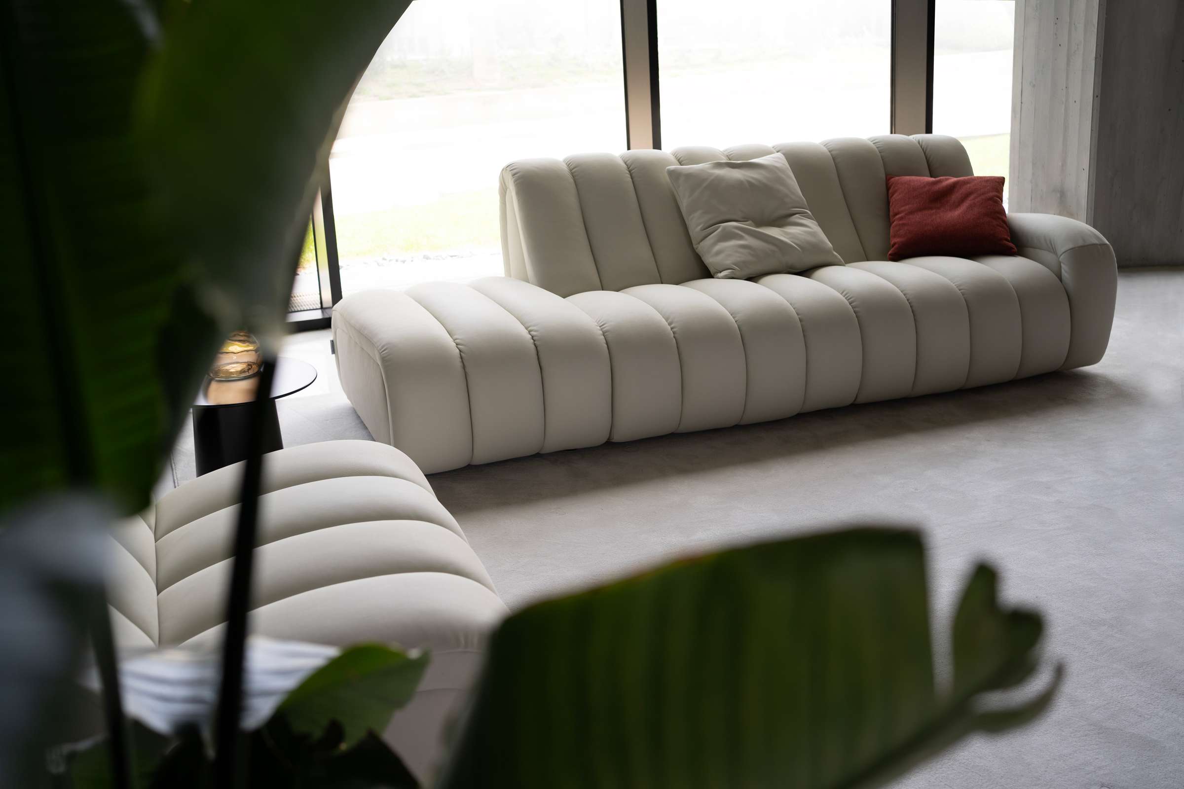 Cream channel-quilted sofa with a red cushion, seen past palm leaves