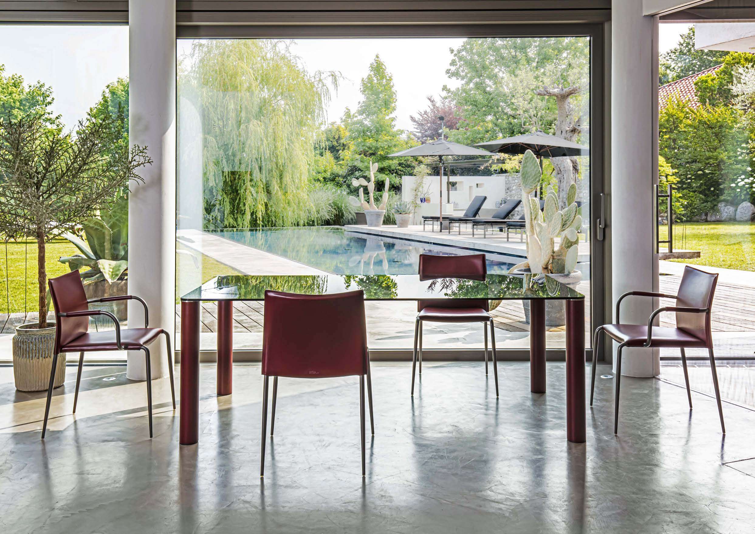 Burgundy leather chairs at a glass table, with a pool and garden beyond the windows