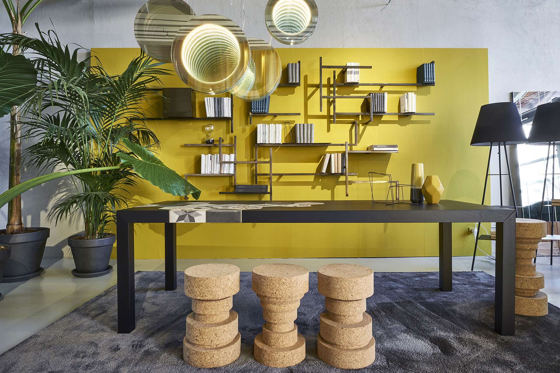 Long dark table with cork stools before yellow wall shelving and round mirrors