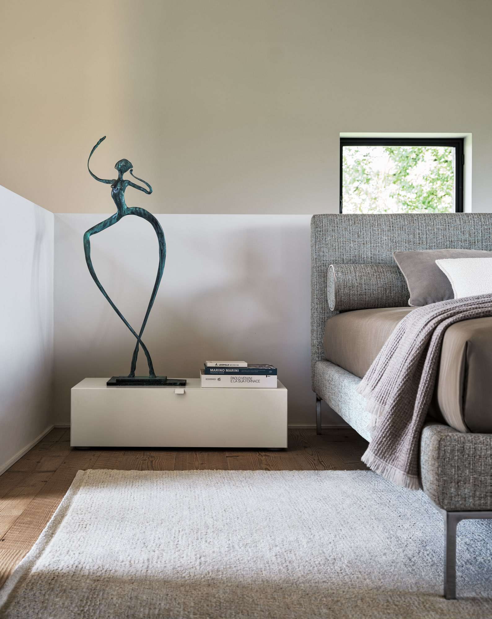 Grey upholstered bed beside a white chest topped with a bronze sculpture