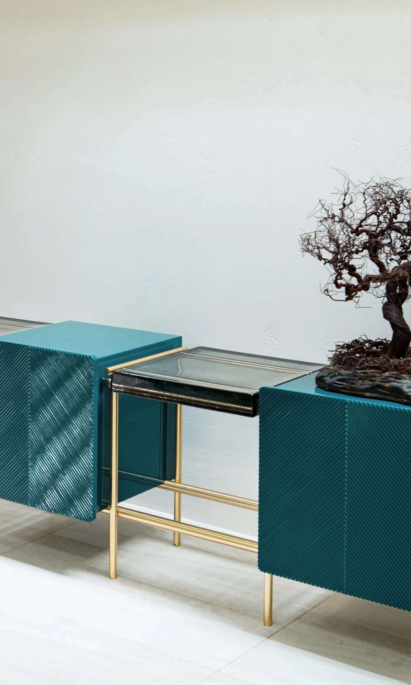 Teal textured storage units with a glass and brass shelf and a bonsai