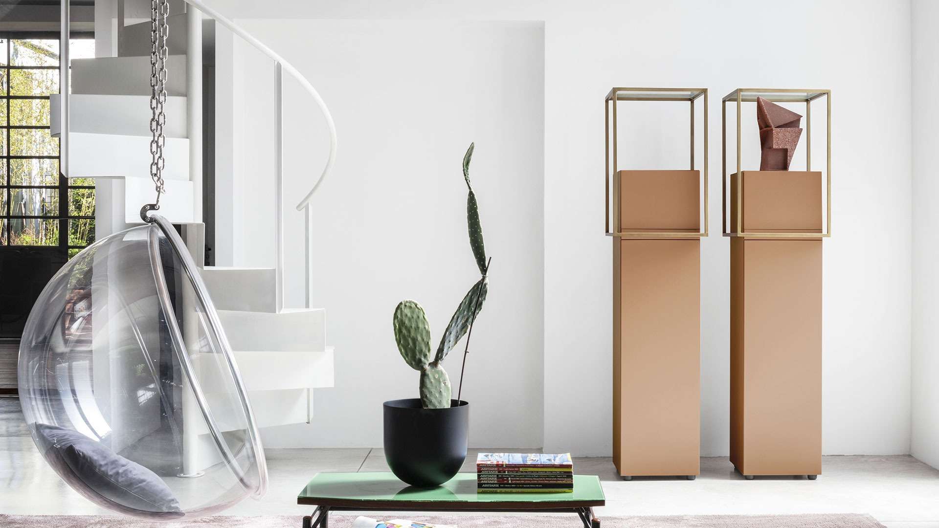 Tall bronze cabinets on plinths and a hanging glass chair in a white room