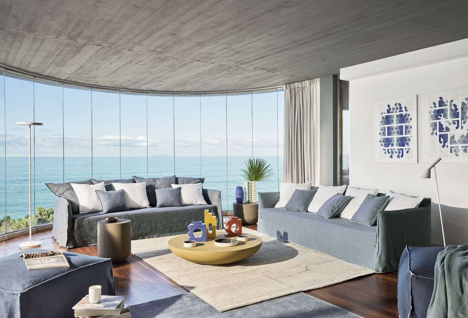 Slipcovered sofas around a yellow coffee table in front of a curved glass wall facing the sea