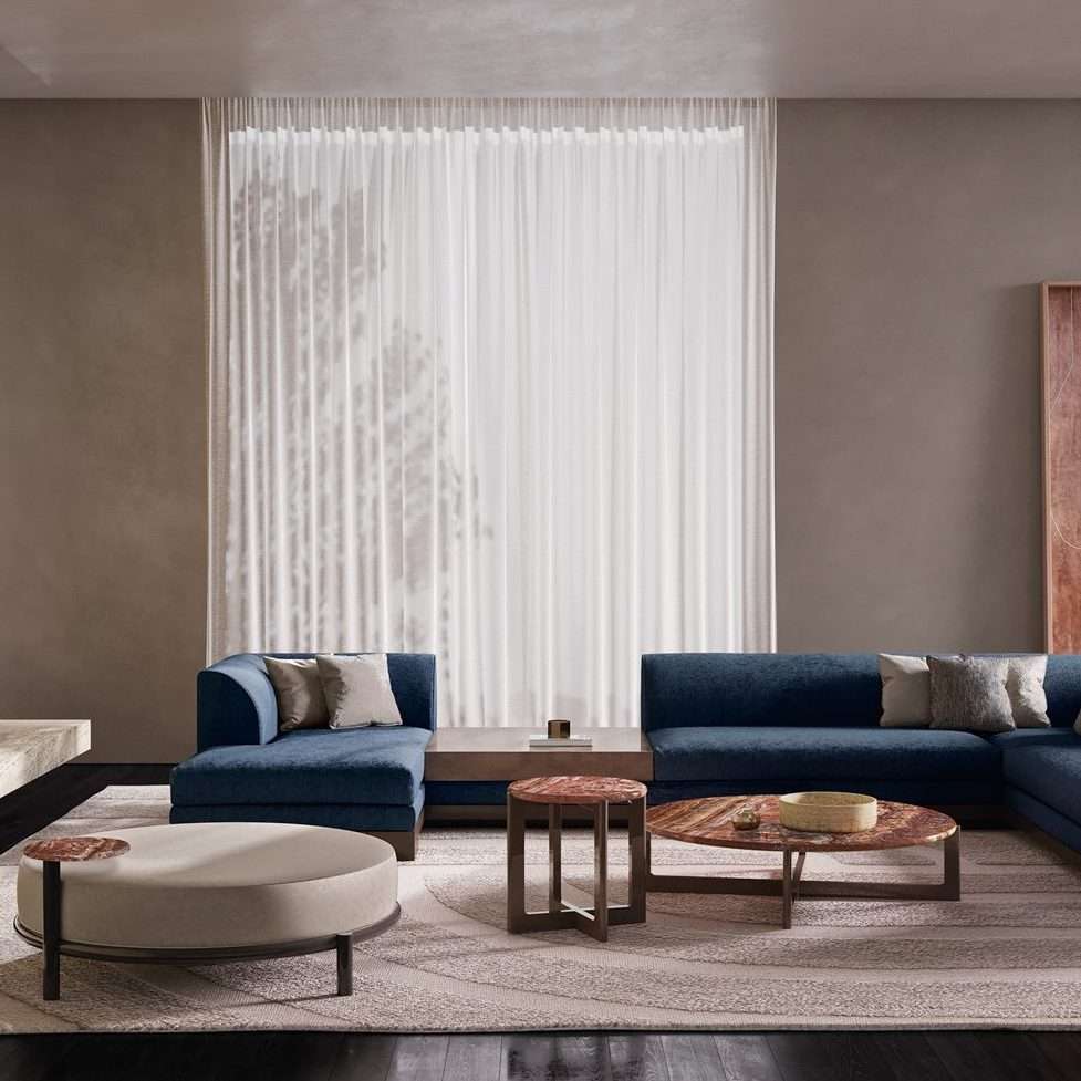 Two blue sectional sofas with a round ottoman and walnut side tables before a sheer curtain