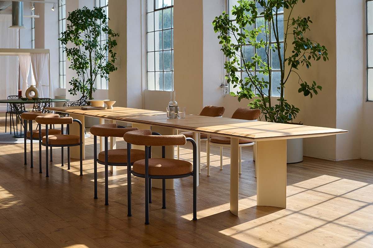 Long pale table and leather chairs in a bright showroom with tall windows and indoor trees
