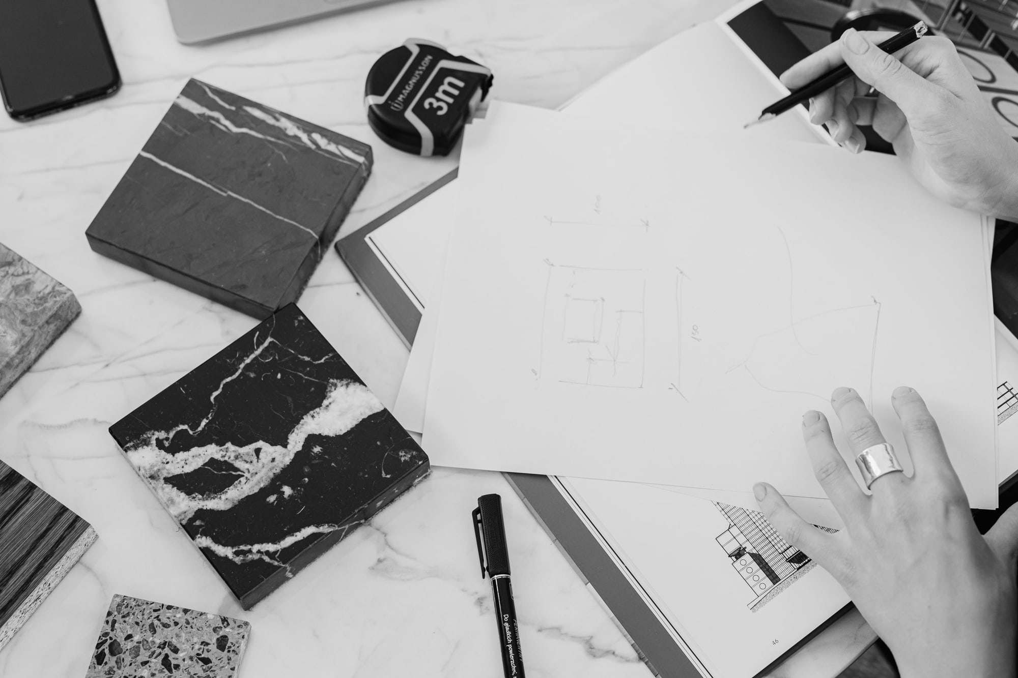 Black-and-white photo of a hand sketching on a plan beside marble samples and a tape measure