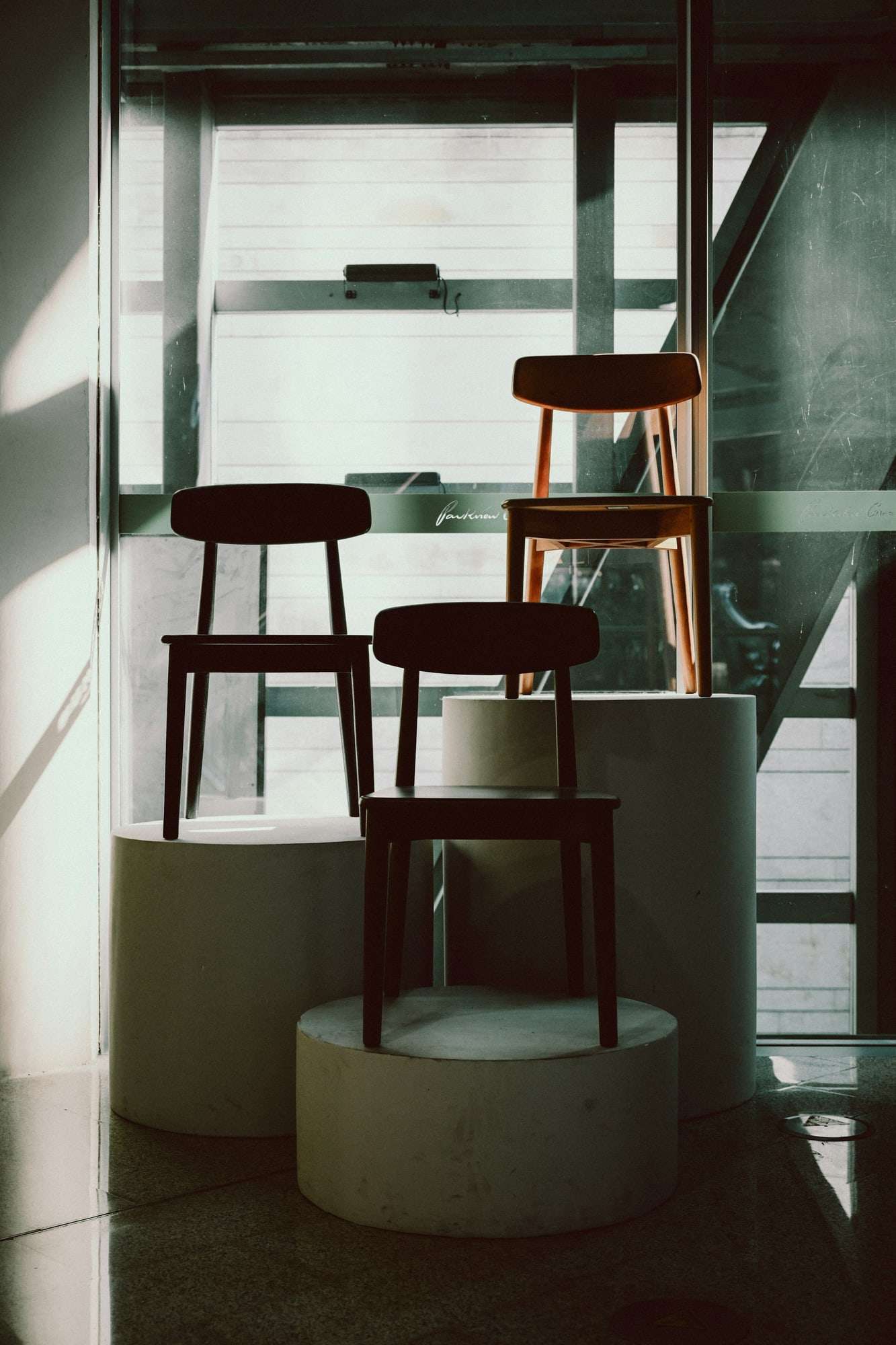 Three wooden chairs on white plinths before a tall window, set out as in a gallery